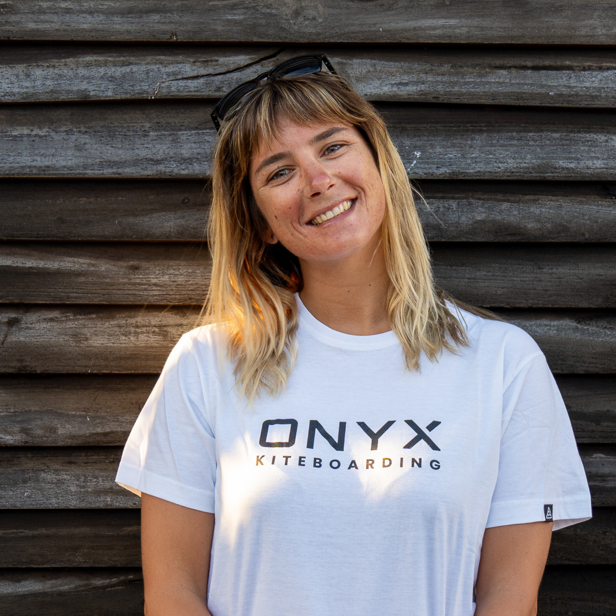 Woman wearing a white 'ONYX KITEBOARDING' t-shirt standing against Wooden wall smiling into camera