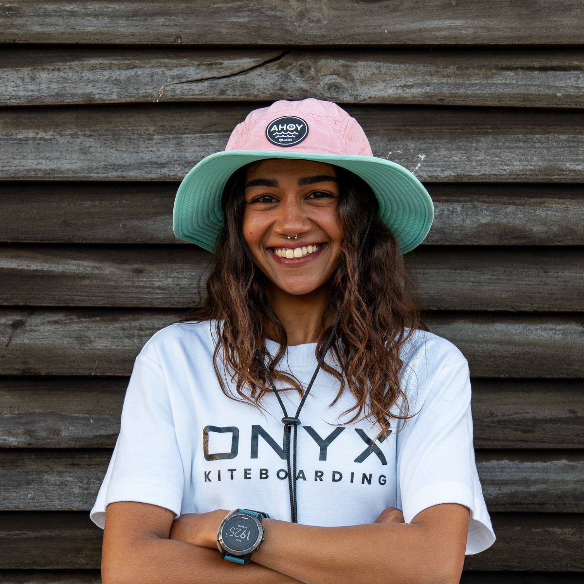 Portrait of woman smiling, wearing a white ONYX Kiteboarding shirt and colorful hat against a wooden background