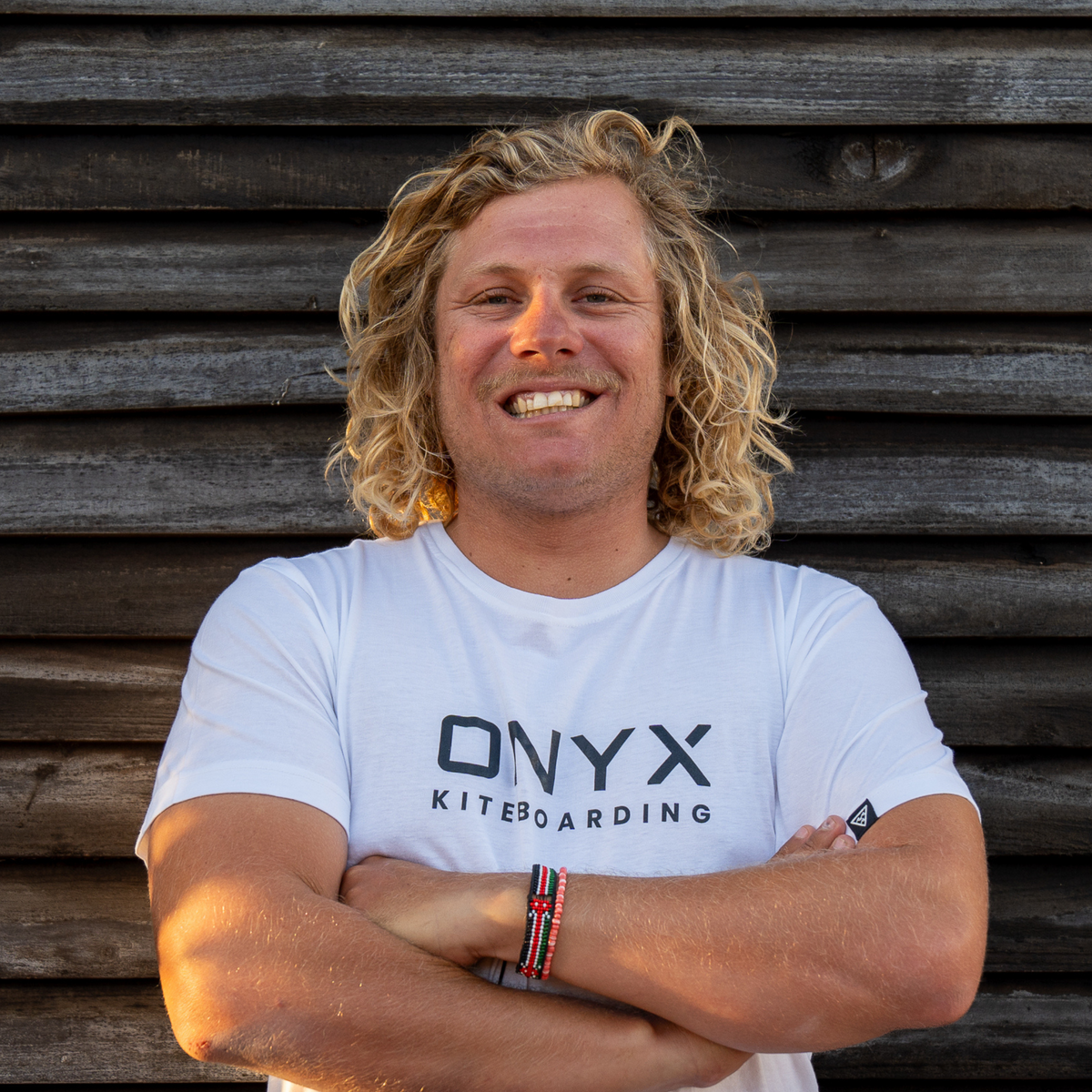 Man wearing an ONYX Kiteboarding t-shirt standing against a wooden wall smiling into camera