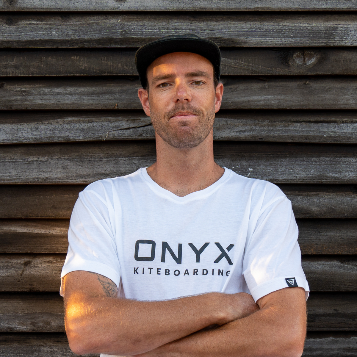 Portrait of a man wearing a white ONYX Kiteboarding t-shirt standing against a wooden wall.
