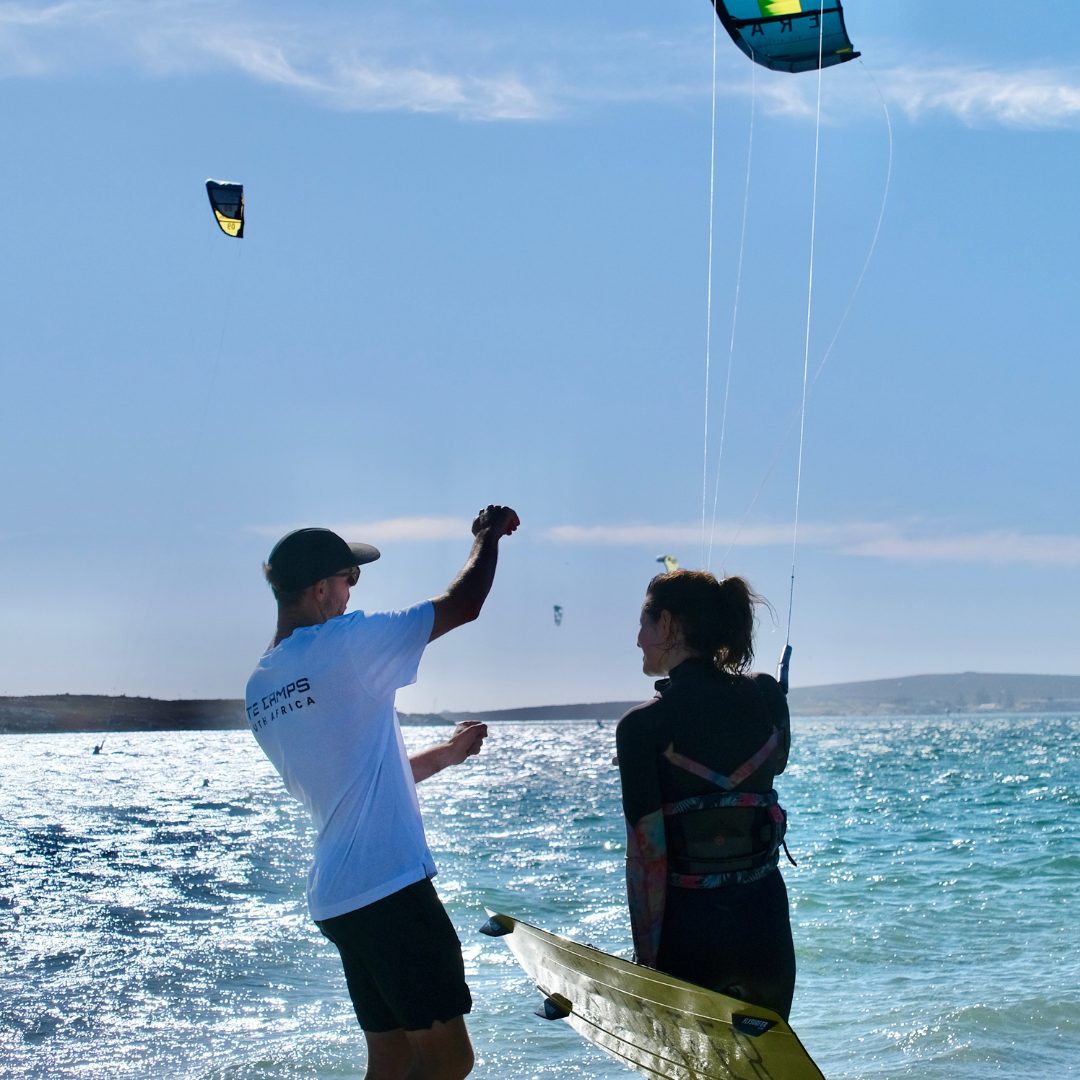 Kitesurfing instructor giving hand signals to a student in a harness at the water's edge in Langebaan Main Beach
