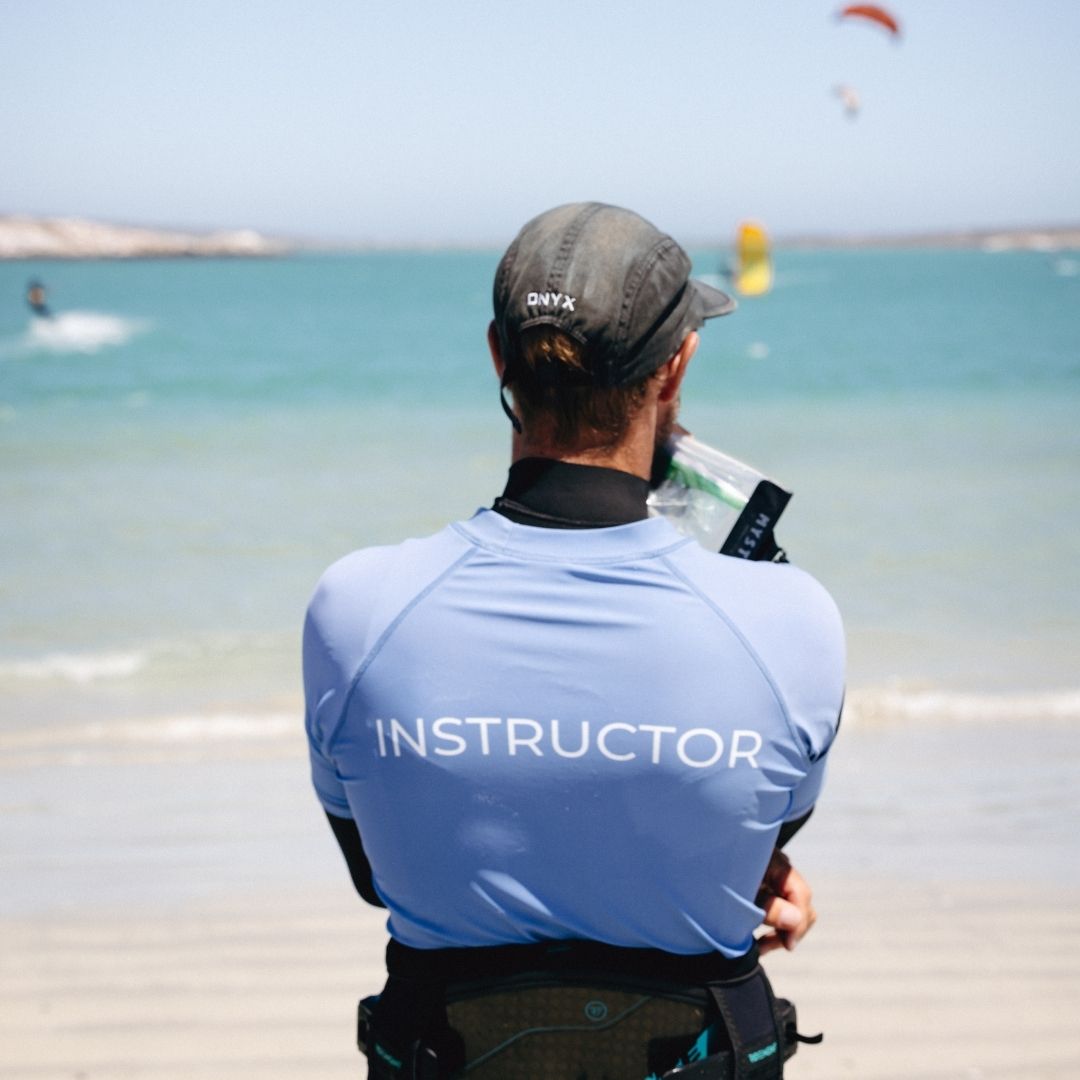 Person wearing a blue 'INSTRUCTOR' shirt on a beach with water and kites in the background talking into a radio